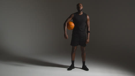 Studioporträt-In-Voller-Länge-Eines-Männlichen-Basketballspielers,-Der-Vor-Dunklem-Hintergrund-Den-Ball-Unter-Dem-Arm-Hält